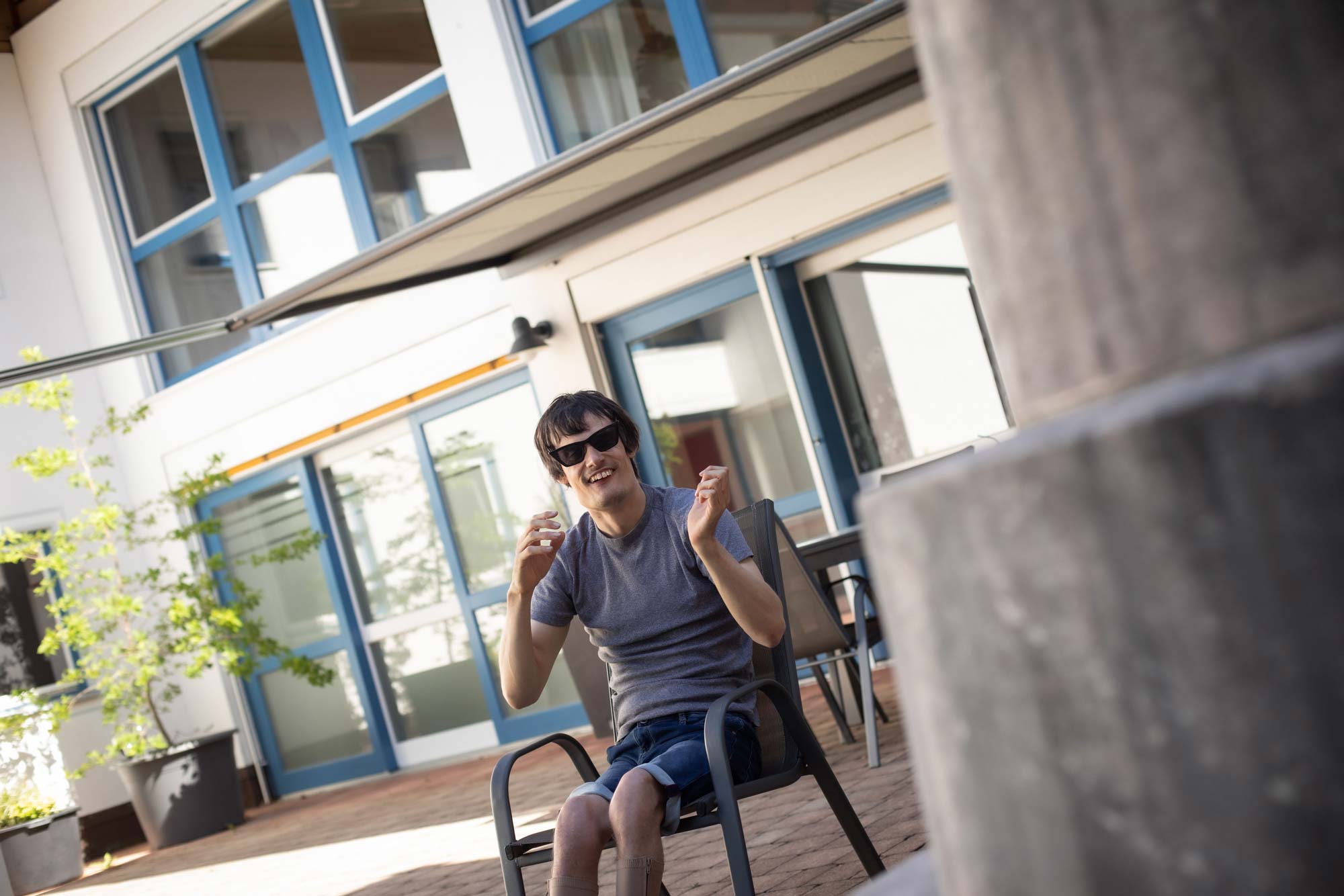 EIn Bewohner sitzt auf der Terrasse in einem Stuhl. Er trägt Sommerkleidung und eine Sonnenbrille und lächelt.