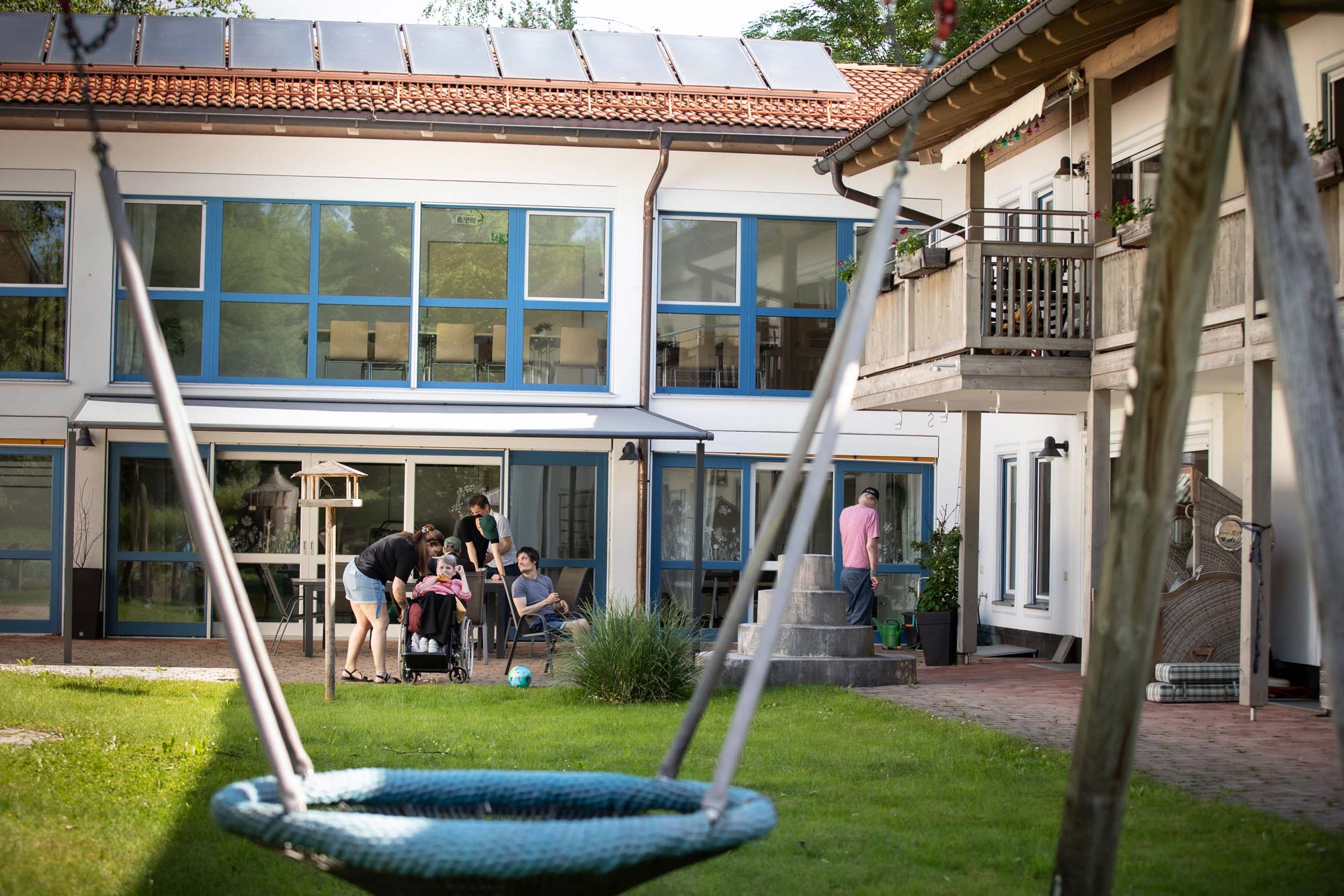 Der Garten des Hauses, mit einer Schaukel. Auf der Terrasse im Hintergrund sind Bewohnerinnen und Bewohner zusammen mit einer Mitarbeiterin zu sehen.
